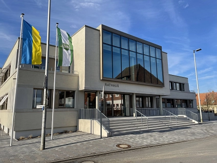 Das sandsteinfarbene Rathaus wird von der Amtsstraße aus gezeigt. Man sieht die große Fensterfront des Sitzungssaals im Obergeschoss und den Haupteingang mit der breiten Treppenanlage sowie dem seitlichen barrierefreien Zugang. Links vom Haupteingang wehe