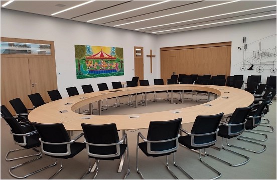 Hier ist der neue Sitzungssaal im kernsanierten Neuen Rathaus abgebildet. In diesem Raum befindet sich ein großer ovaler Sitzungstisch mit schwarzen Stühlen für dreißig Personen.
