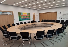 Hier ist der neue Sitzungssaal im kernsanierten Neuen Rathaus abgebildet. In diesem Raum befindet sich ein großer ovaler Sitzungstisch mit schwarzen Stühlen für dreißig Personen.
